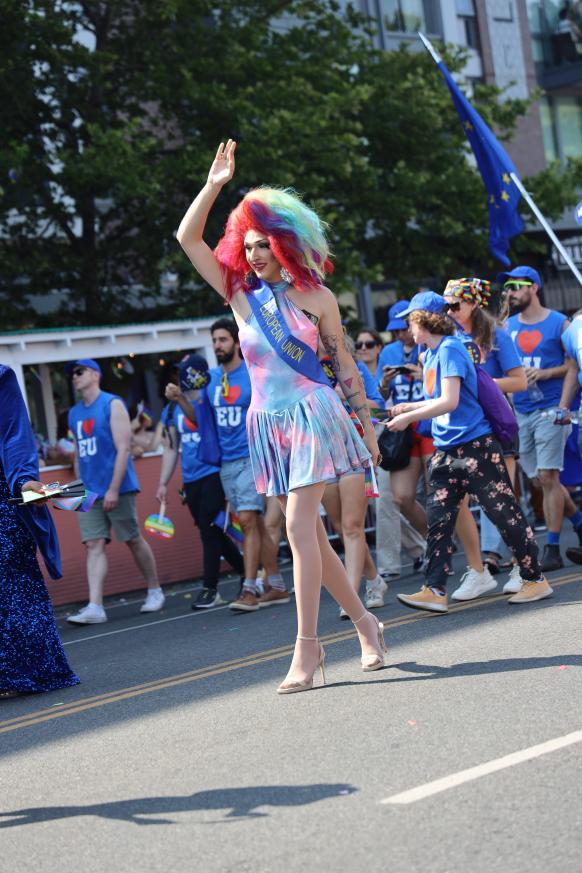 A drag queen in a rainbow wig waves at parade onlookers.