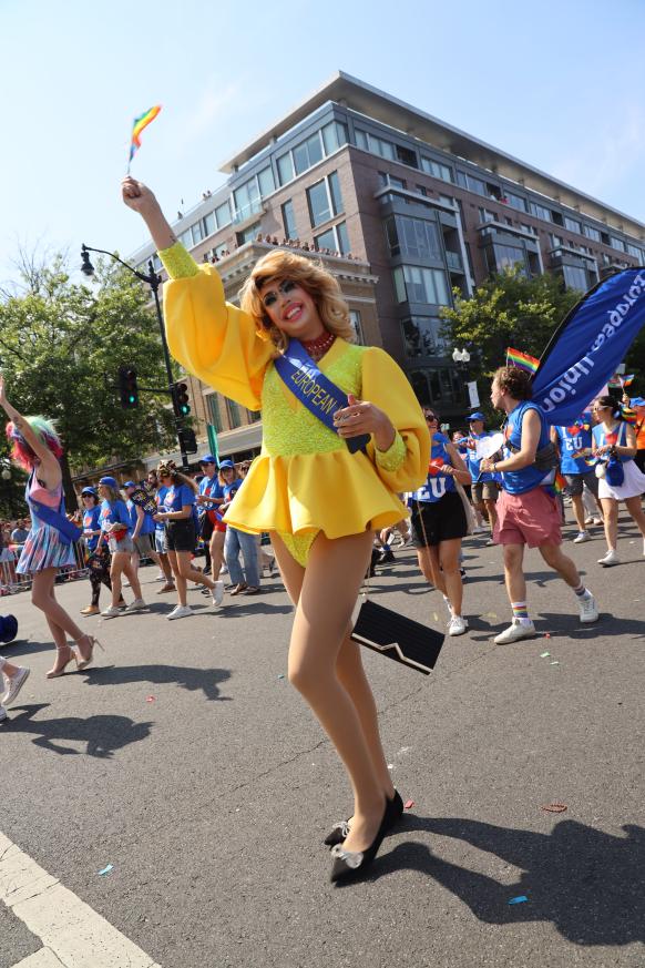 A drag queen in a yellow dress waves a Pride flag.