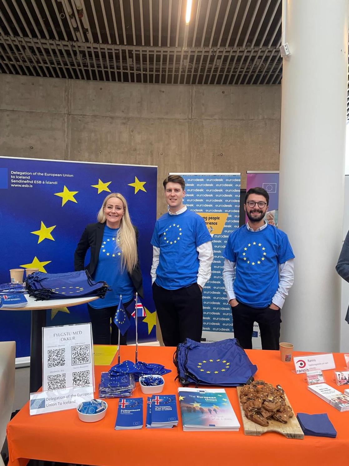 three people standing in a booth decorated with EU flags and promotional material 