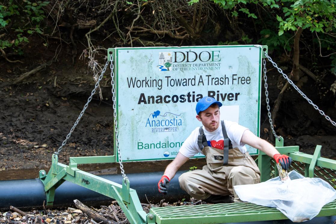 A young man picks up trash in front of a sign that says "Working Toward a Trash Free Anacostia River."