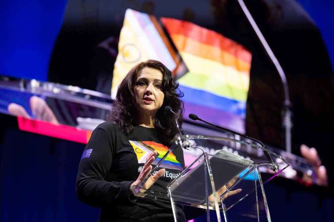 Person in pride t-shirt at lectern delivering a speech