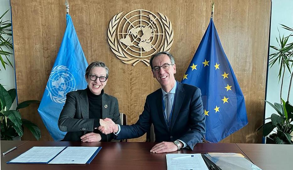 Woman and man shake hand with UN and EU flags in background.