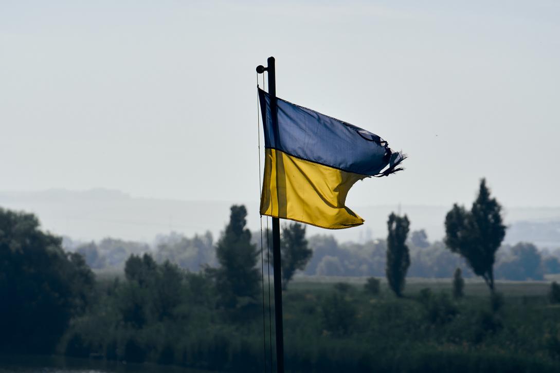 Tattered Ukraininan flag flies above a battlefield on a grey day.